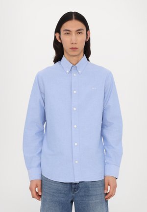 Light blue button-up shirt made of woven fabric, featuring a collar, long sleeves, and white buttons. Subtle branding on the chest.