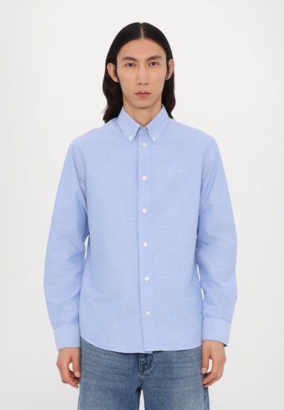 Chemise boutonnée bleu clair en tissu tissé, dotée d'un col, de manches longues et de boutons blancs. Marquage subtil sur la poitrine.