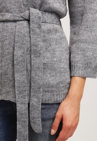 Šedý pletený kardigan s měkkou strukturou, který má v pase zavazovací pásek a volné rukávy. Na viditelném lemu a manžetách jsou viditelné žebrované detaily.