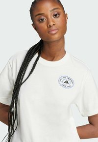 White cotton t-shirt featuring a round logo in blue and black on the left chest, with short sleeves and a relaxed fit.