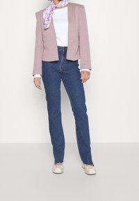 Kvinna klädd i en rosa blazer, vit topp, blå jeans med hög midja, krämfärgade klackar och en lila halsduk med prästkragar, stående mot en vit vägg.