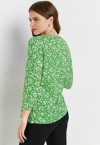 Grünes Oberteil mit floralem Muster, dreiviertel Ärmeln, rundem Ausschnitt und lockerem Schnitt. Das Material wirkt leicht und hat eine glatte Textur.