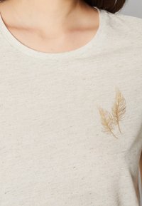Camiseta de algodón beis claro con cuello redondo, que presenta un sutil diseño de hoja dorada en el lado izquierdo. Tela texturizada con un patrón moteado.