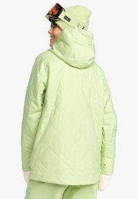 Svetlozelená prešívaná bunda s kapucňou, ktorá má voľný strih a detailné prešitie. Outfit dopĺňa jemná zelená čiapka a lyžiarske okuliare.