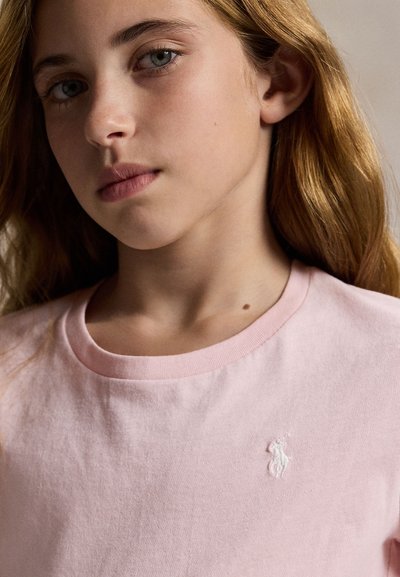 Persoană tânără cu păr lung castaniu deschis, purtând un tricou roz pal cu un mic logo alb brodat pe piept.