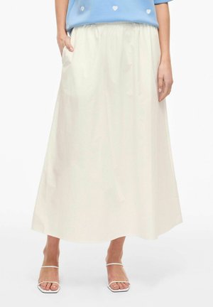 Femme portant une jupe crème longue jusqu'aux chevilles avec des poches latérales et des sandales blanches à brides, assortie à un haut bleu avec des détails en forme de cœur.