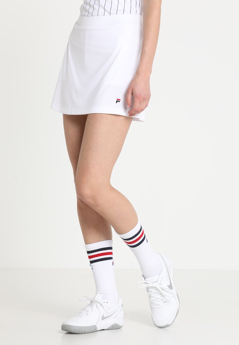 Jupe de tennis blanche avec une taille ajustée, fabriquée en tissu lisse. Associée à des baskets blanches et à des chaussettes mi-mollet rayées. Logo Fila visible.