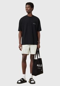 Black oversized t-shirt, light cream shorts, black sandals, white socks; holding a black tote bag with text, standing against a gray background.