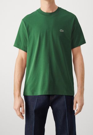 Mężczyzna w prostym zielonym t-shircie z krótkim rękawem z małym logo krokodyla na piersi i ciemnoniebieskich jeansach, stojący na jednolitym tle.