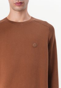 Rjava pletena jopica z okroglim izrezom, rebrasti detajli in majhna reliefna logotip na prsih. Gladka tekstura in prilega oblikovanju.