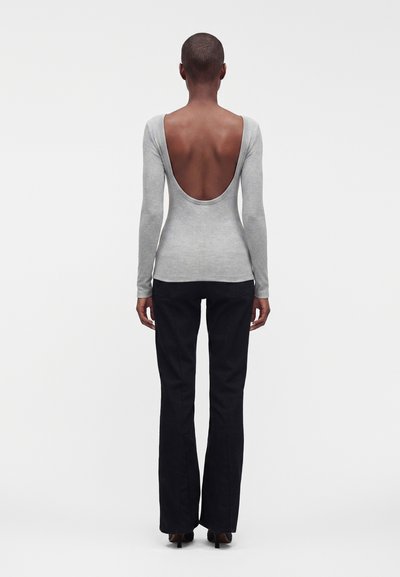 Personne debout de dos portant un haut gris clair à manches longues avec un profond décolleté dans le dos et un pantalon noir avec des talons.