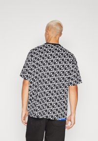 Svart kortärmad skjorta med ett upprepat vitt logomönster. Rak skärning med rund hals och en blå logopatch på nederkantens vänstra sida.