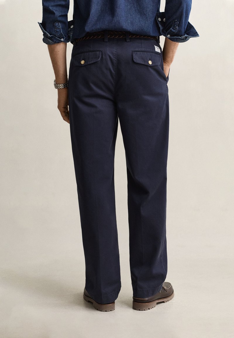 Persona che indossa pantaloni blu navy con tasche posteriori con bottoni, scarpe marrone scuro e una camicia di jeans a maniche lunghe arrotolata, vista di spalle.