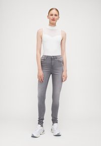 Valkoinen hihaton bodysuit, jossa on muotoiltu kaula-aukko, yhdistettynä harmaisiin skinny-farkkuihin. Mallilla on valkoiset lenkkarit. Yksinkertainen tausta.