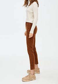 Femme portant un pull en maille torsadée blanc, un pantalon en velours côtelé marron et des baskets épaisses beiges, debout contre un fond clair uni.