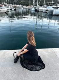 Black lace skirt with a floral pattern and solid black top, seated on a concrete dock beside a marina, holding a black handbag.