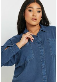 Camicia di denim con bottone frontale, due tasche sul petto e maniche arrotolate. Il colore è blu medio con una leggera texture e una vestibilità rilassata.