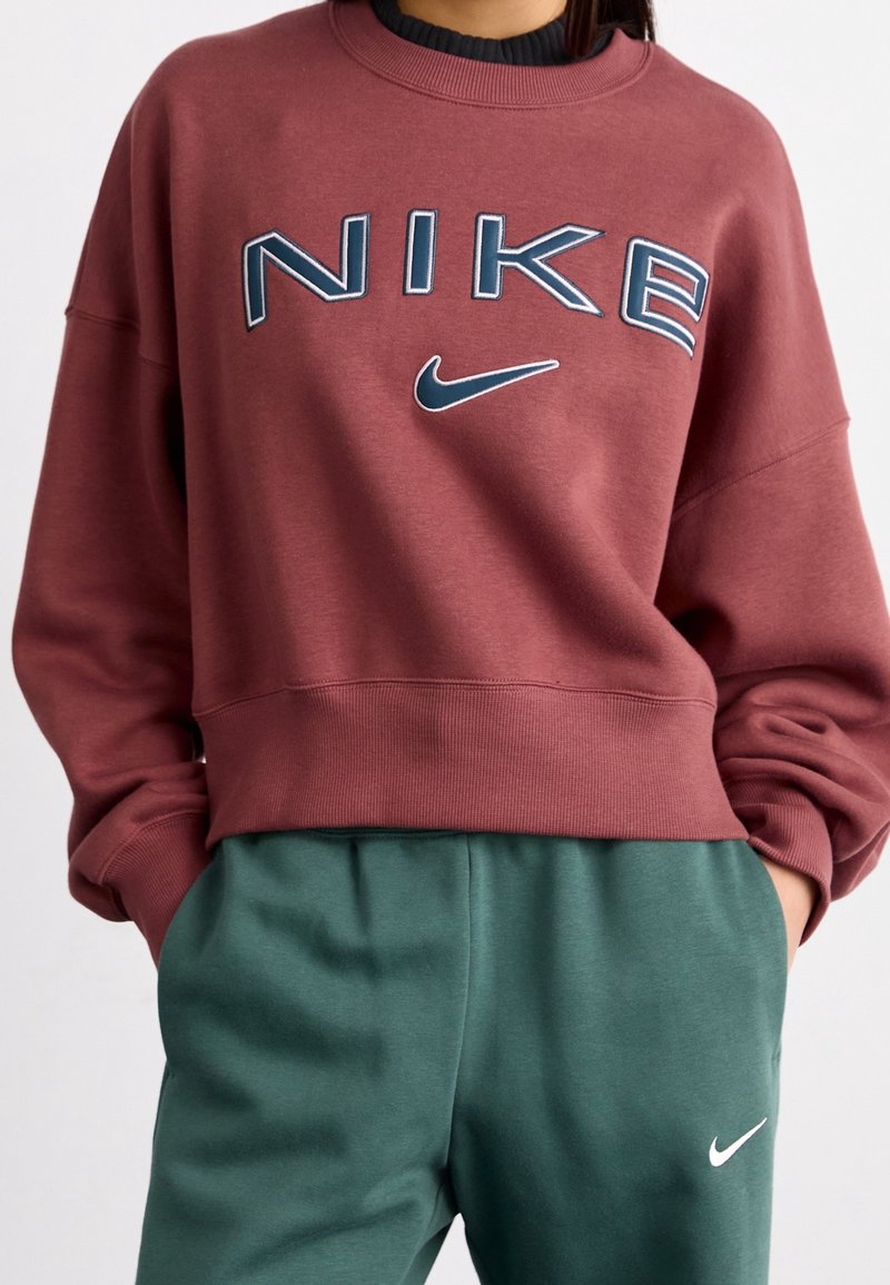 Maroon huppari, jossa on "NIKE"-logo sinisenä ja valkoisena. Ominaisuuksiin kuuluu miehustan muotoinen kaula-aukko ja löysä istuvuus. Yhdistetty tummanvihreisiin jumppahousuihin.