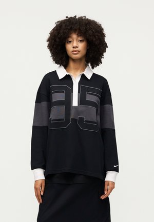 Mujer con cabello rizado que lleva una camiseta de manga larga negra y gris con cuello y puños blancos, con un gran "95" en el frente.