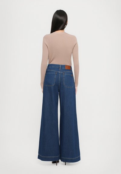 Laiad sinised denim teksad kõrge vöökohaga, kahe tagataskuga ja pruuni nahkpatchiga vöökohtade peal. Sile tekstuur, vabaja disain.