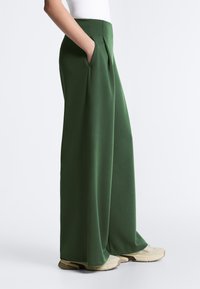 Persoană purtând pantaloni verzi cu talie înaltă și croială largă, cu mâna în buzunar, asortați cu adidași bej și un top alb, pe un fond simplu.