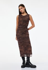 Frau in einem ärmellosen, braunen Midikleid mit Tiermuster und schwarzen, klobigen Stiefeln, steht mit einer Hand hinter ihrem Rücken.