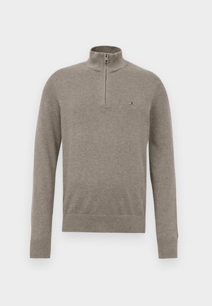 Svetra svetlo rjave barve s polovičnim zadrgo, dolgimi rokavi, rebrastimi manšetami in robom ter majhnim logotipom na levem prsnem delu.