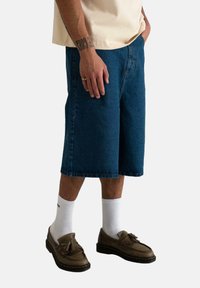 Shorts en denim bleu à coupe ample, de conception longueur genoux, avec poches avant, portés avec des chaussettes blanches et des mocassins marron. Texture douce, look décontracté.