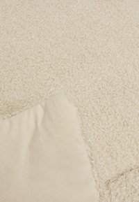 Tapis texturé beige montré partiellement avec une surface douce et pelucheuse ; côté présentant un bord en tissu léger et lisse.