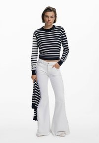 Navy- und weiß gestreifter Strickpullover mit langen Ärmeln, kombiniert mit weißen, ausgestellten Jeans. Das Model hält einen passenden gestreiften Schal.