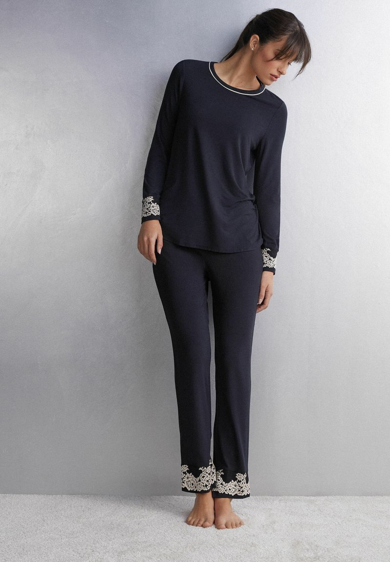 Marineblauw pyjama-set van zachte stof, met lange mouwen en een broek met delicate witte kanten accenten bij de manchetten en de zoom.