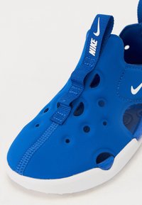 Blauer Sportschuh aus flexiblem Material, mit nahtlosem Design, Belüftungsperforationen und einem weißen Logo, ausgestattet mit einer Schlaufenriemen.