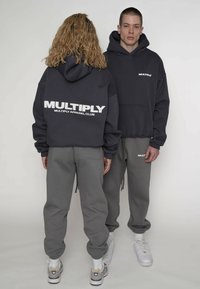 Hanorac gri închis cu glugă și logo-ul "MULTIPLY" pe spate; asortat cu pantaloni de trening gri deschis și adidași albi. Material texturat.