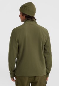  giacca in pile verde oliva con colletto alto, maniche lunghe e superficie liscia. Abbinata a pantaloni cargo verdi oliva e un berretto.