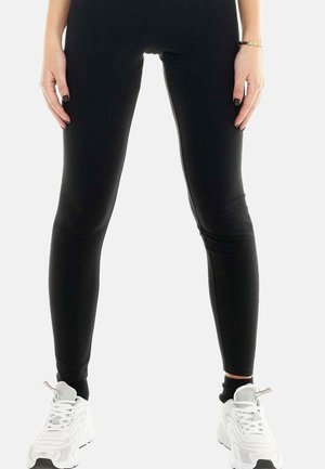 Vrouw draagt strakke zwarte legging en witte sportieve sneakers, staand met ontspannen handen langs zij op een effen witte achtergrond.