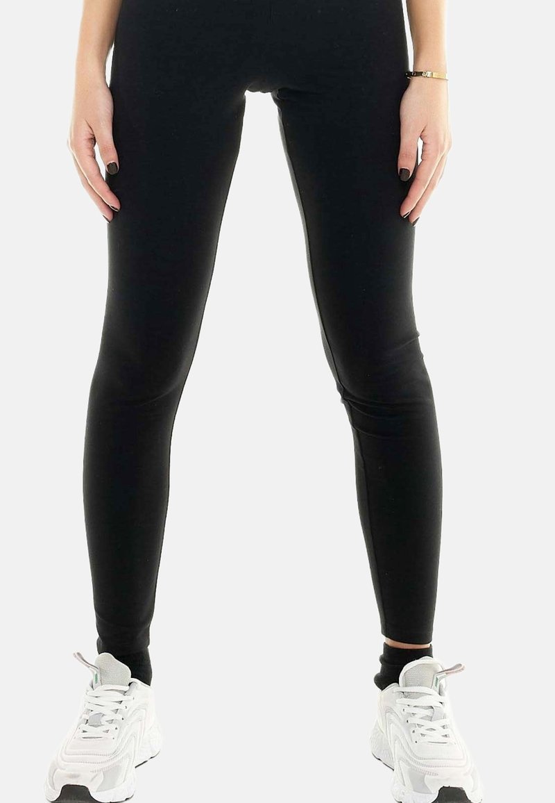 Femme portant des leggings noirs moulants et des baskets blanches de sport, debout les mains détendues le long du corps sur un fond blanc uni.