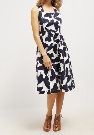 Vrouw draagt een witte mouwloze midi-jurk met grote zwarte vlinderprints, gecombineerd met bruine open-toe veterlaarzen tot aan de enkels.