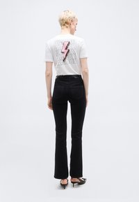 Άτομο με κοντά ξανθά μαλλιά, φορώντας λευκό μπλουζάκι με ροζ αστραπή και το κείμενο «AMOUR», μαύρο τζιν και μαύρες ψηλοτάκουνες, γυρισμένο πλάτη.