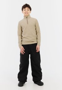 Beige rits fleece top met hoge kraag, gecombineerd met losse zwarte sneeuwbroeken en donkere schoenen, staande tegen een witte achtergrond.