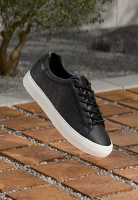 Sneaker in pelle nera caratterizzati da un motivo testurizzato, punta rotonda, suola in gomma bianca e lacci neri, posizionati sopra una superficie piastrellata.