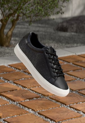 Sneaker in pelle nera caratterizzati da un motivo testurizzato, punta rotonda, suola in gomma bianca e lacci neri, posizionati sopra una superficie piastrellata.
