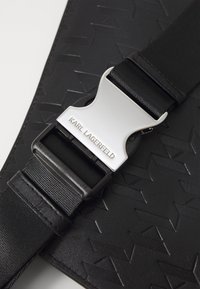 Fekete texturált szövet, mintázattal, ezüst színű fémből készült csat található rajta, "KARL LAGERFELD" felirattal gravírozva, egy fekete állítható pánthoz rögzítve.