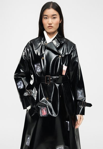 Γυναίκα που φοράει μία γυαλιστερή μαύρη καμπαρντίνα με ζώνη και πολλούς μικρούς φωτογραφικούς επιθέματα, όρθια μπροστά σε απλό ανοιχτό φόντο.