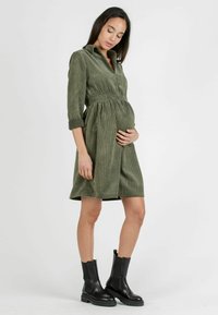 Olijfgroene corduroy jurk met een kraag, elastische tailleband, driekwartmouwen en een uitlopende rok. Gestyled met zwarte enkellaarzen.