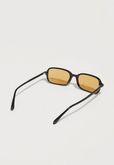 Μαύρα ορθογώνια γυαλιά ηλίου με λεπτά πλαϊνά και φακούς με κεχριμπαρένια απόχρωση, ανοιχτά και τοποθετημένα σε λευκή επιφάνεια.