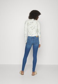 Camiseta de manga larga blanca con estampados florales, combinada con unos jeans ajustados azules y zapatos de lona beige, vista desde la parte posterior.