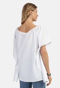 Blusa bianca ampia con scollatura larga, maniche corte e fessure laterali, realizzata in tessuto leggero, mostrata da dietro.