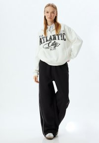 Witte sweatshirt met "ATLANTIC CITY" in zwart gedrukt, gecombineerd met losse zwarte broek met witte zijkanten en grijze sneakers.