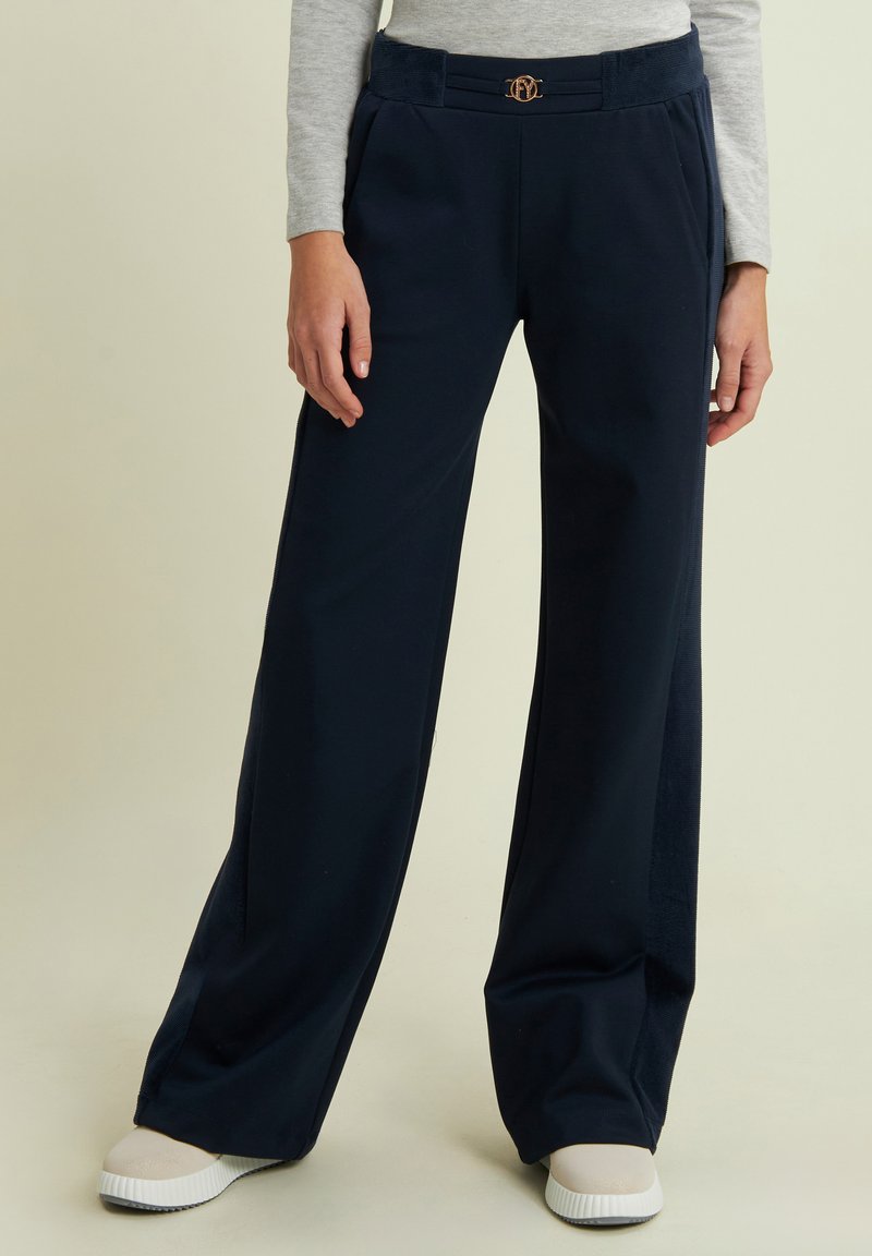 Pantaloni a gamba larga blu navy realizzati in un tessuto morbido, con una vita testurizzata e un dettaglio logo metallico sul davanti. Completati da sneakers bianche.