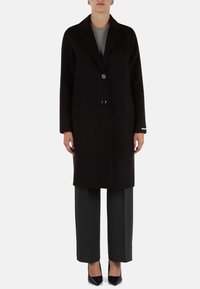 Cappotto di lana nera con chiusura a un pulsante, revers a punta e due tasche laterali, abbinato a pantaloni grigi sartoriali e tacchi neri.